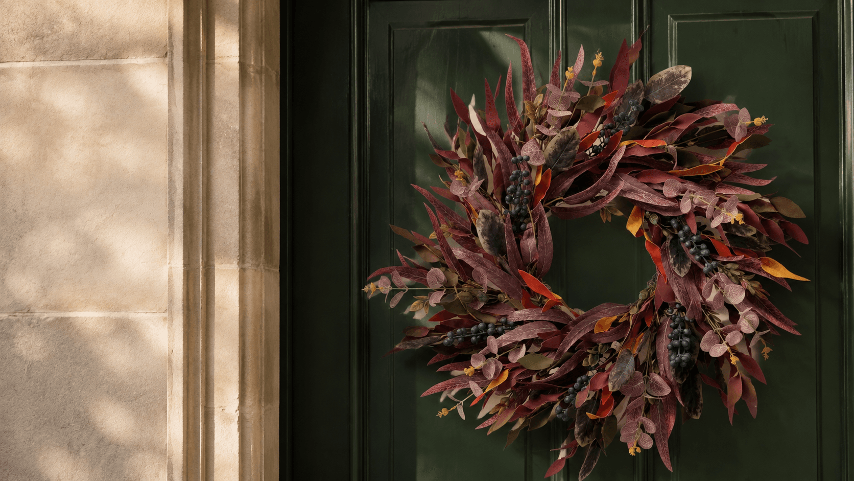 A burgundy eucalyptus and blueberry wreath on a forest green front door in warm autumn light